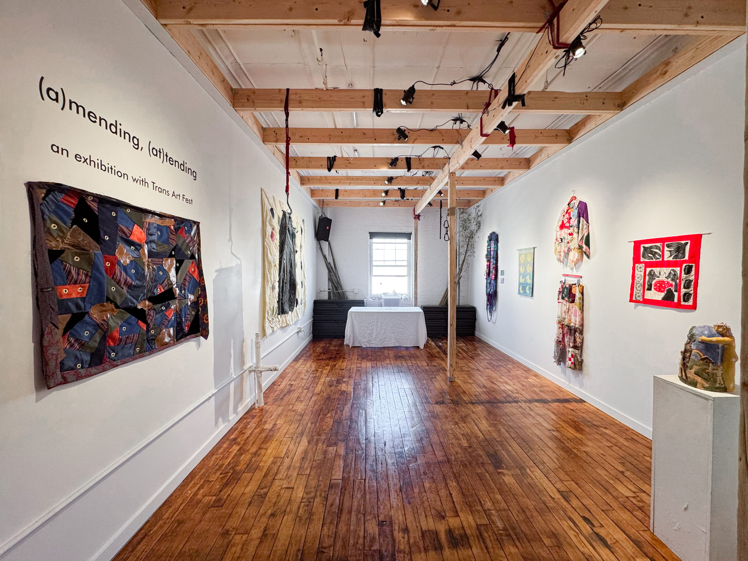Installation view of Coil & Cord’s white-walled gallery space with warm wood floors, exposed beam ceilings, and track lighting. Works on view include textiles and garments hung along the walls, with a ceramic sculpture on a pedestal in the foreground.​​​​​​​​​​​​​​​​